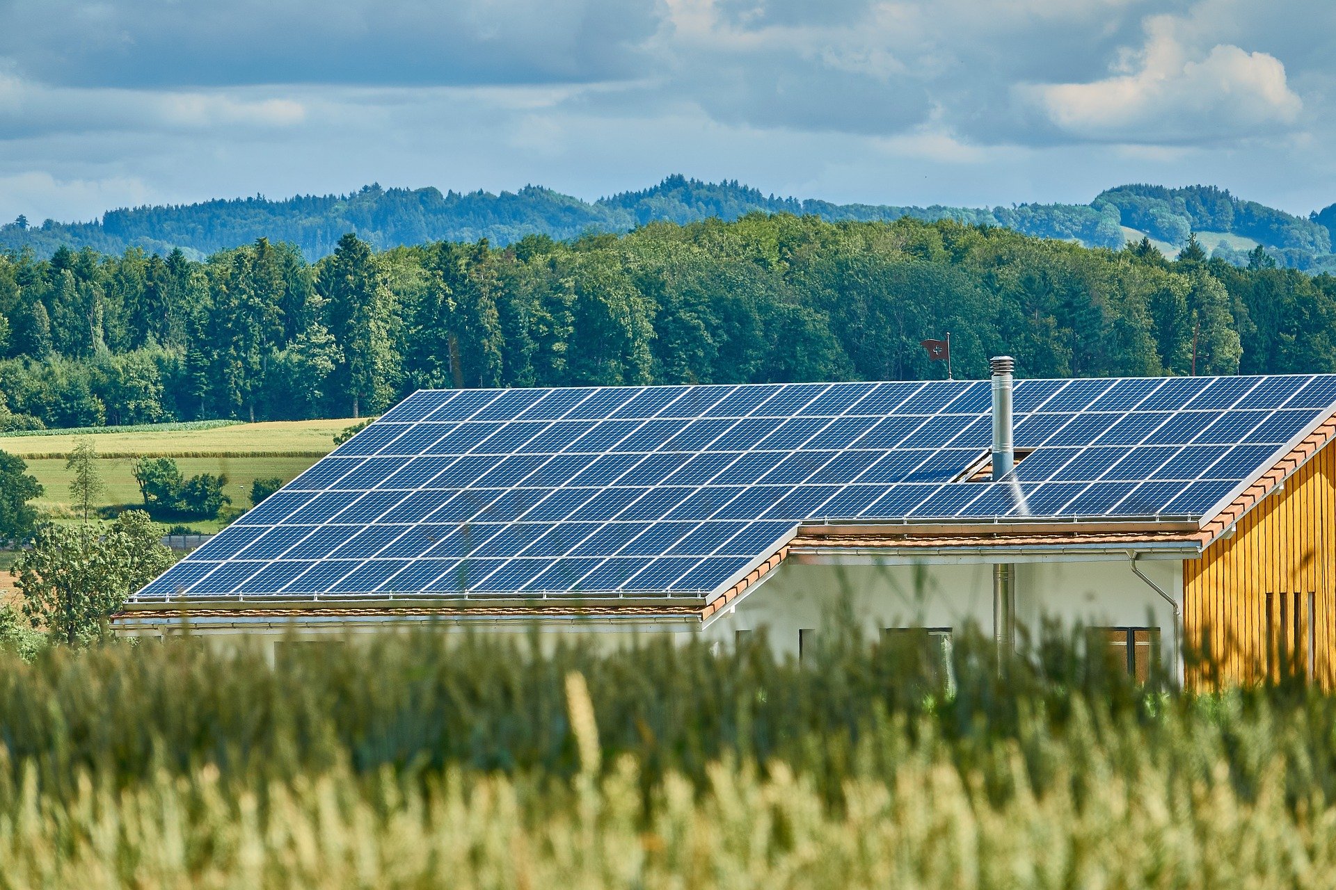 Ein Gebäude mit einer Dachfläche als Photovoltaikanlage vor Feldern und im Hintergrund Wald